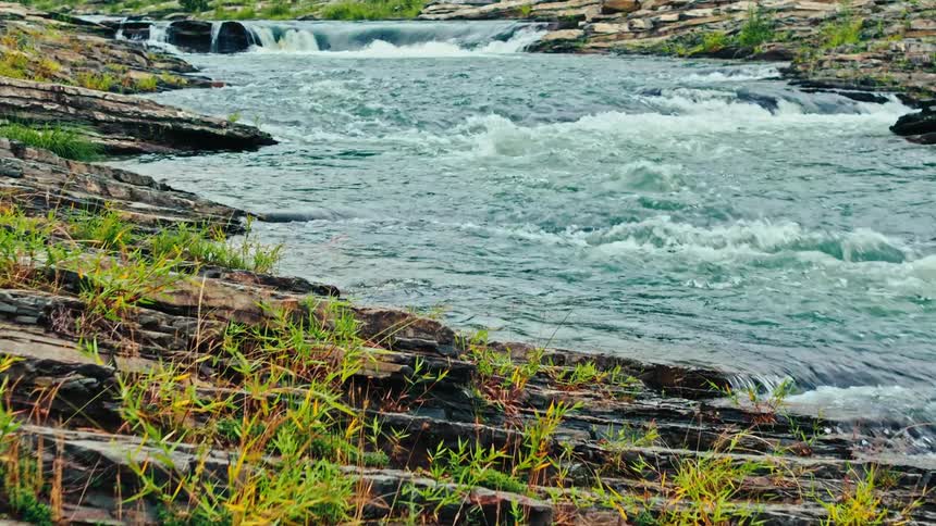 Sedimentary rocks with sparse vegetation in the foreground and a river with a small waterfall in the background