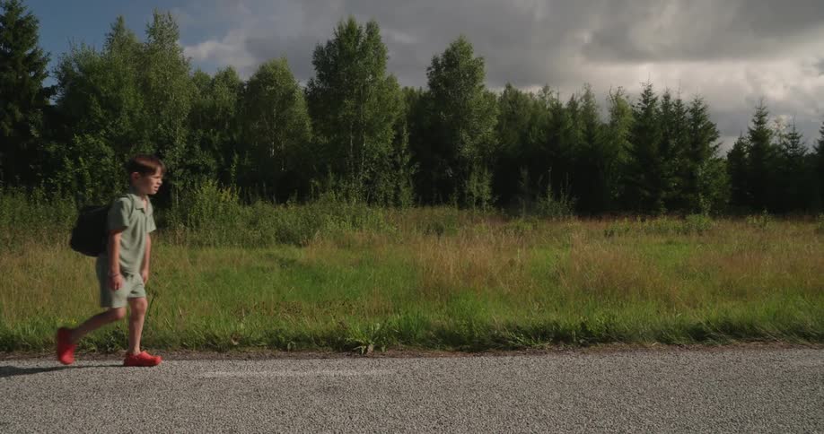A child with a backpack walking on a dirt road beside a forest