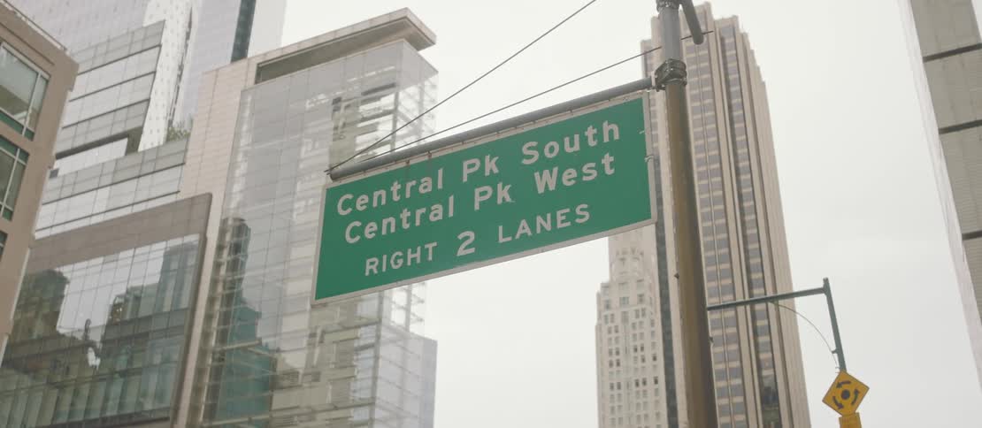 A green traffic sign with white writing and behind it huge skyscraper buildings