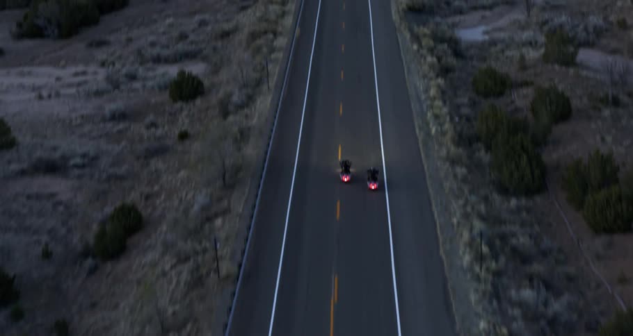 Two motorcycles traveling along a paved road surrounded by a desert environment with low vegetation