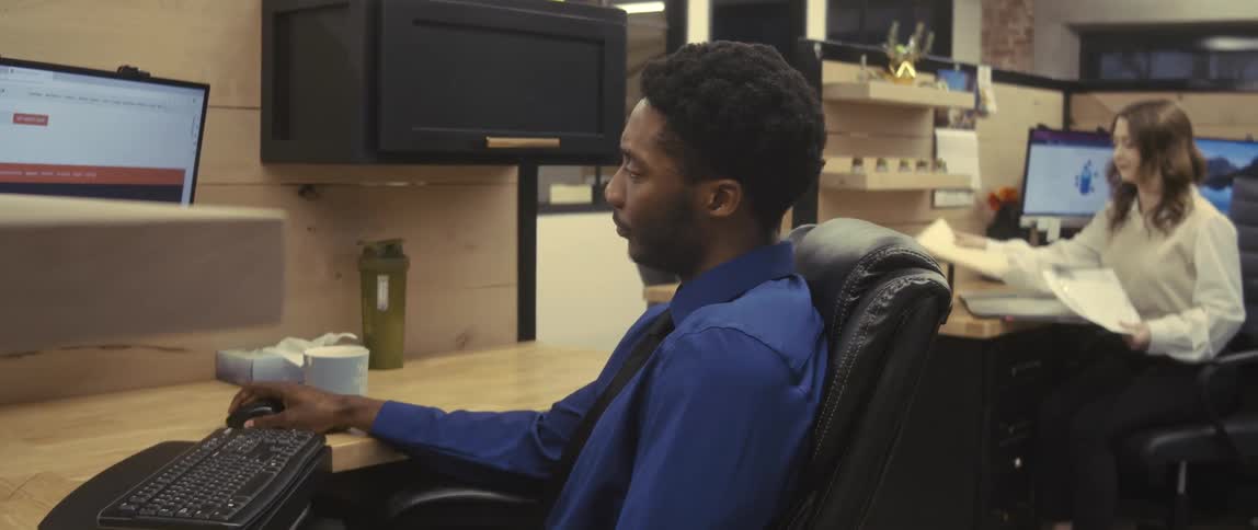 A young man in a blue shirt is sitting and shaking his head while looking at a computer and then starts typing on the keyboard
