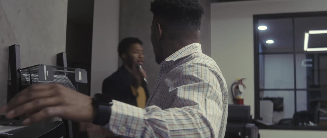A man in a checkered shirt standing beside a printer holding a sheet of paper while processing documents in an office setting