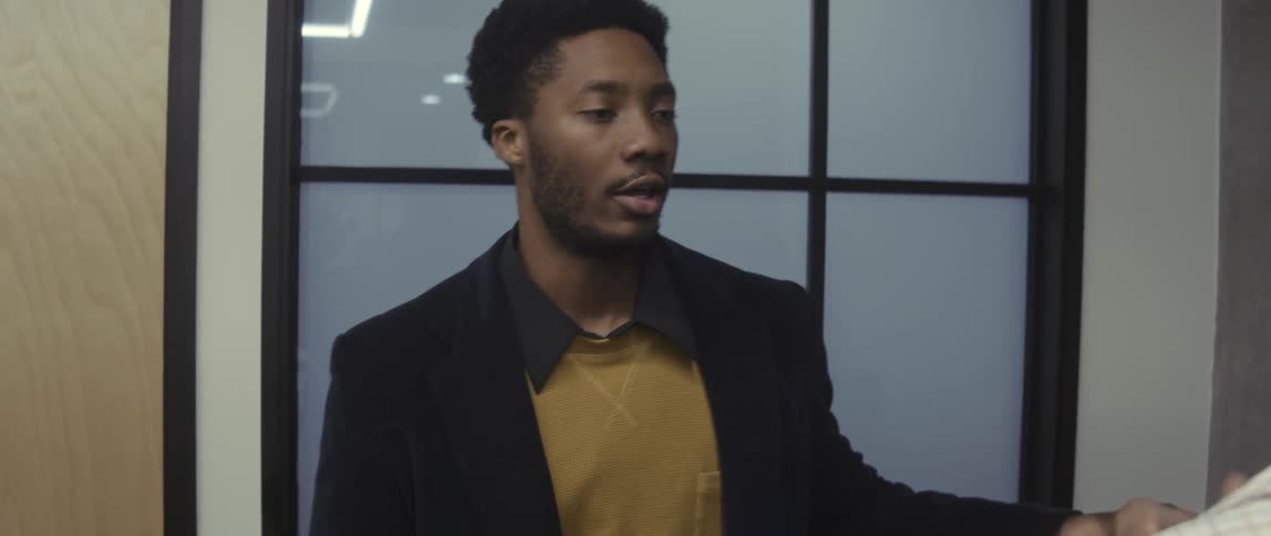 A male employee walks down a hallway and speaks with another employee in an office