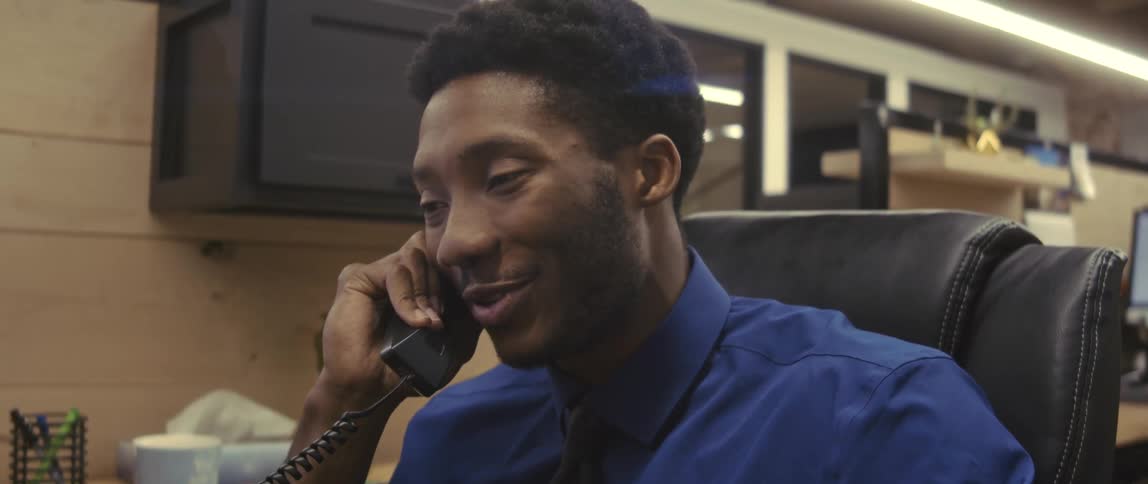 A young man sitting in an office chair wearing a blue dress shirt is talking on the phone and he is smiling