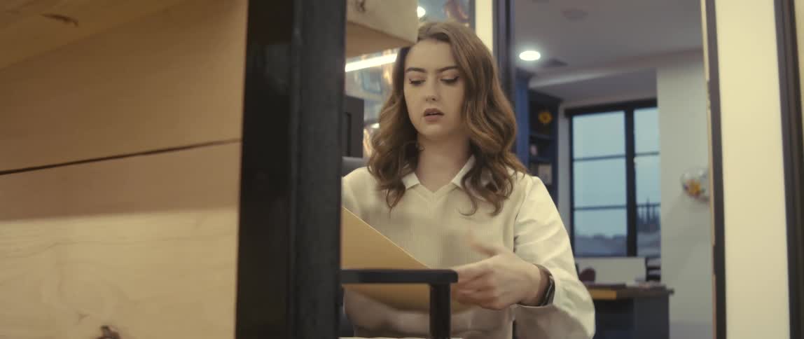 A young woman in an office with a paper in her hand and then she opens it and starts reading it