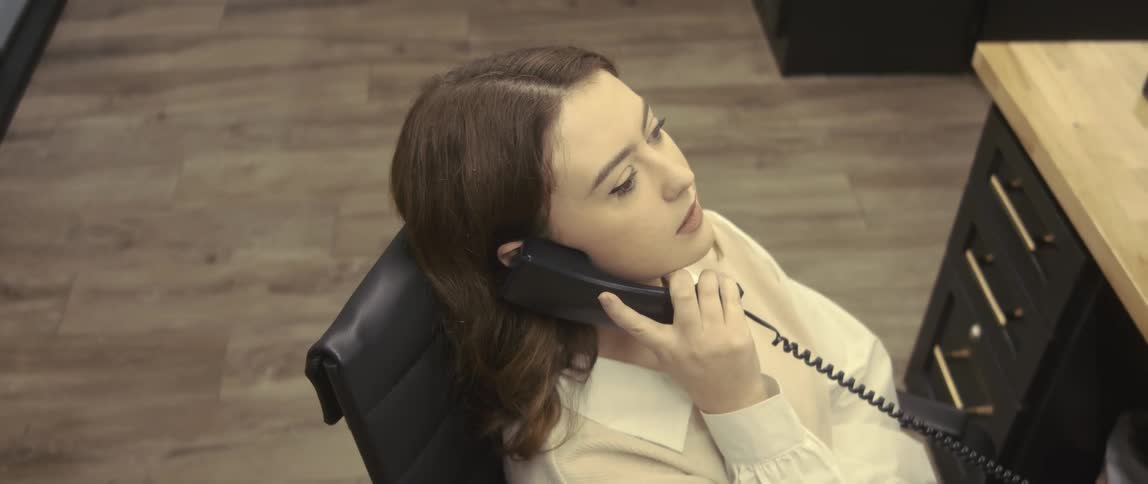 A young woman is sitting in an office chair and has a phone to her ear and she starts to smile