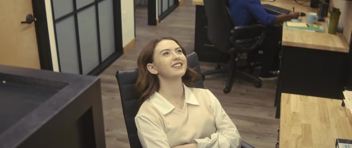A white female employee leans back in her chair and smiles as a male coworker sits at a desk nearby