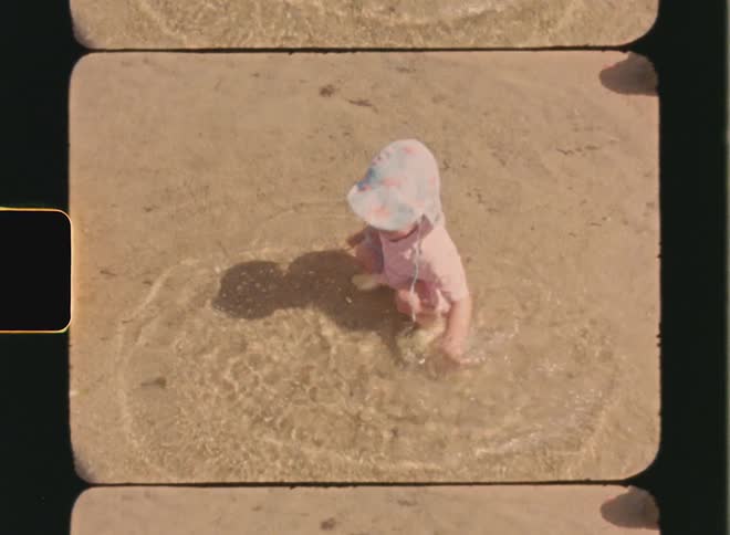 Super 8 film of a little girl in a hat. She is squatting in the sea and she is playing with her arm in the water