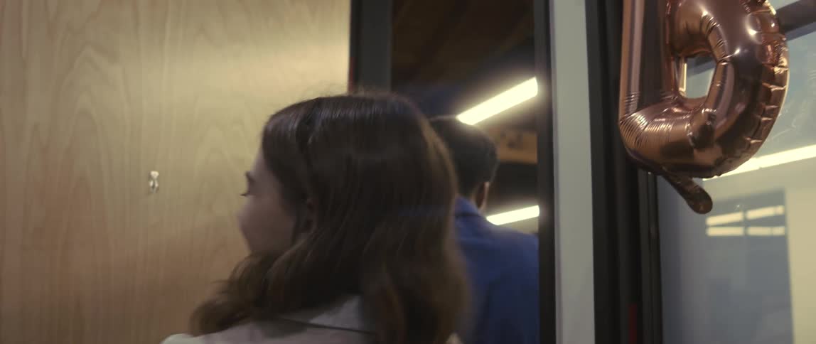 A young woman and a young man at an office birthday party smile and nod as they leave the room closing the door behind them