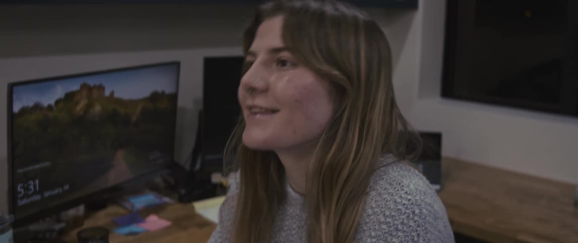A young woman sits at an office workspace while smiling and talking