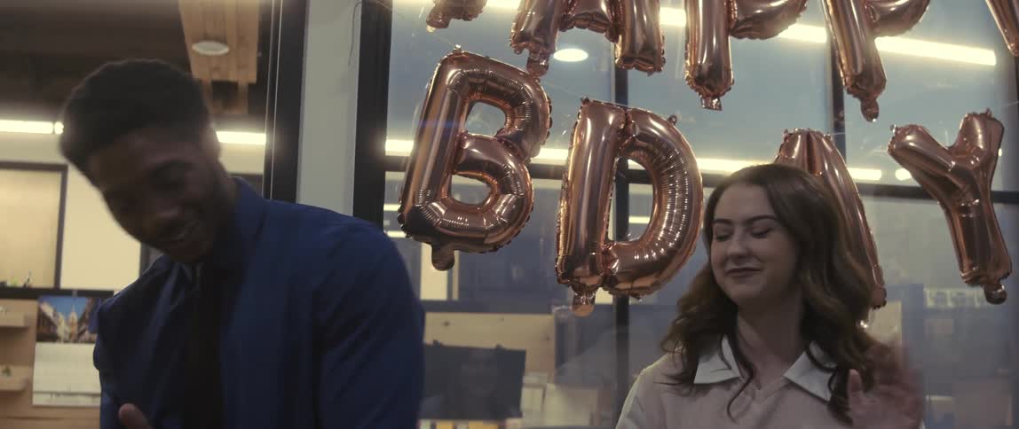 A male and female employee smile and wave before leaving an office with birthday balloons hanging on a window