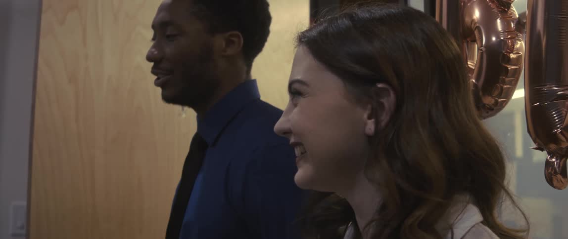 A man and a woman are in office clothes and happy while talking to someone who is in front of them and next to them there are some balloons