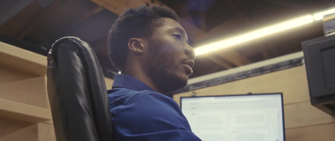 An office worker wearing a blue shirt talking to someone while sitting on his cubicle