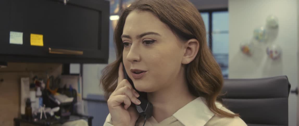 Businesswoman with brown hair and a white shirt sits at desk and talks on the phone at the office