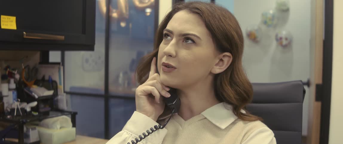 A businesswoman with brown hair wearing a shirt sits in an armchair and talks on the phone in her office