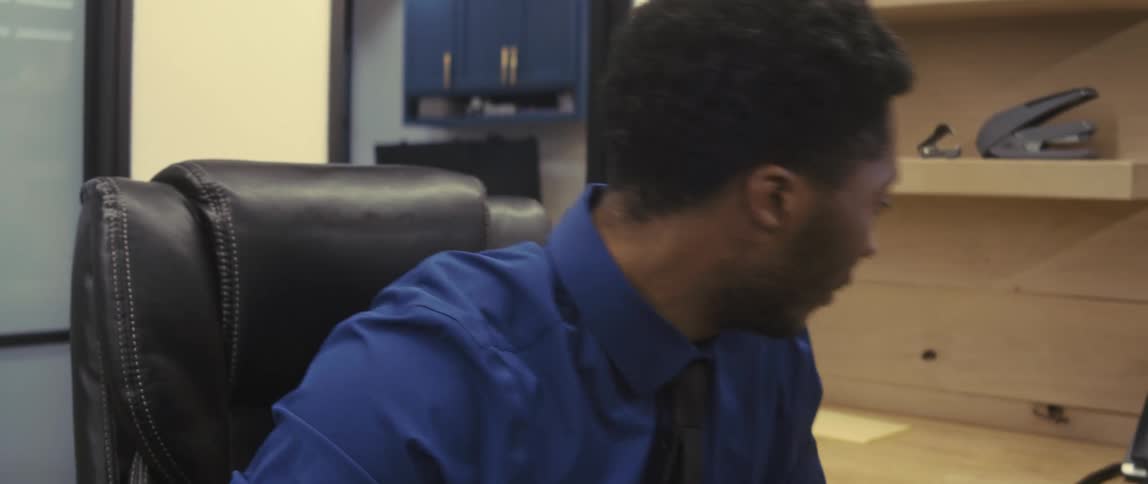 A businessman dressed in a blue collared shirt and a black tie is sitting in a black leather office chair nodding and works on computer