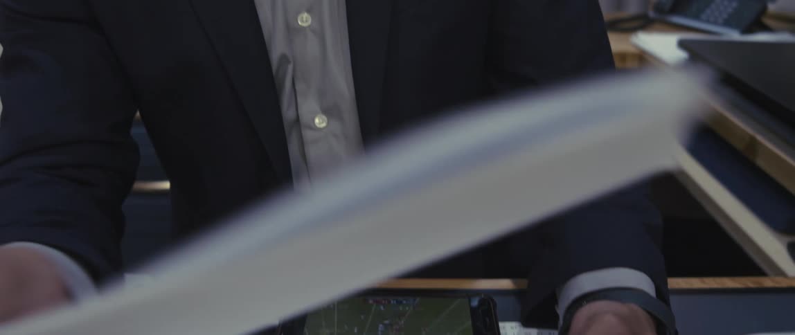 Man sitting at an office desk and checking documents while watching a game on his phone