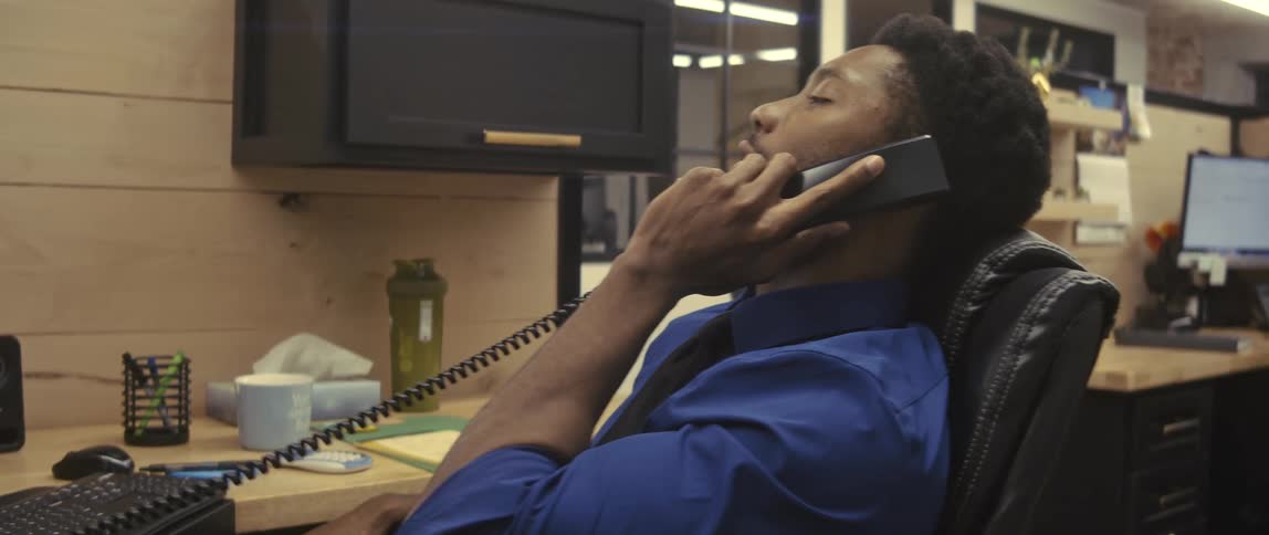A businessman dressed in a blue collared shirt and a black tie is sitting in a black leather office chair talking on a phone at the office