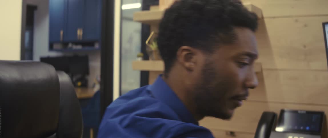 A young man in a blue shirt is sitting in an office chair looking to his right side and then he looks forward