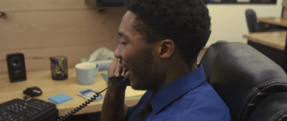A young man in an office cubicle talks on the phone and laughs