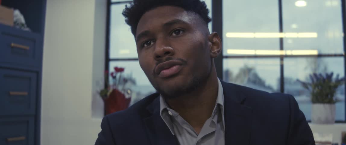 A person in a dark suit sits indoors with a glass window in the background