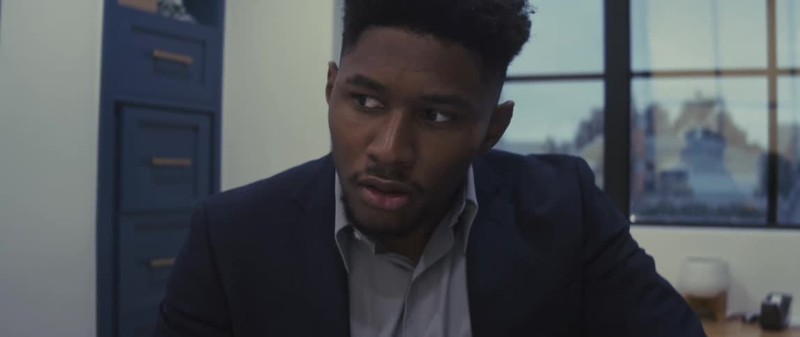 A young man in formal clothes is sitting looking straight ahead and then he starts thinking and puts his hand on his head