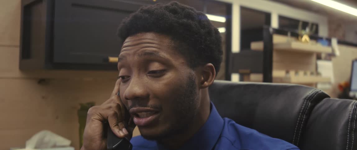 A young man in an office cubicle talks on the phone then looks over his shoulder