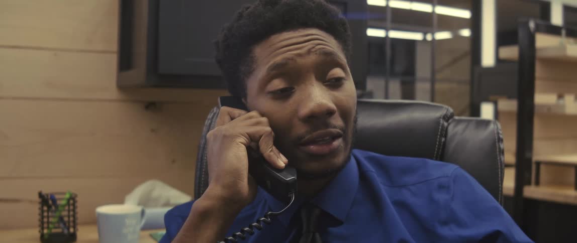 A man in a blue shirt is talking on the phone and he is sitting in an office and he is nodding his head in affirmative tones