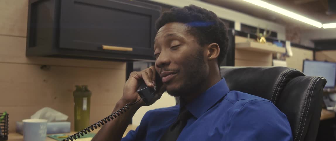 A businessman wearing a black tie and a blue shirt sits in a leather chair and talks on the phone in the office