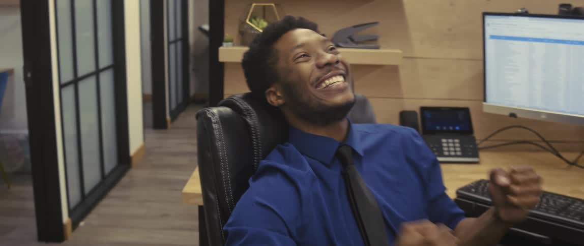 A man in a blue shirt and black tie sitting in an office chair laughing joyfully celebrating or feeling excited while leaning back in his seat