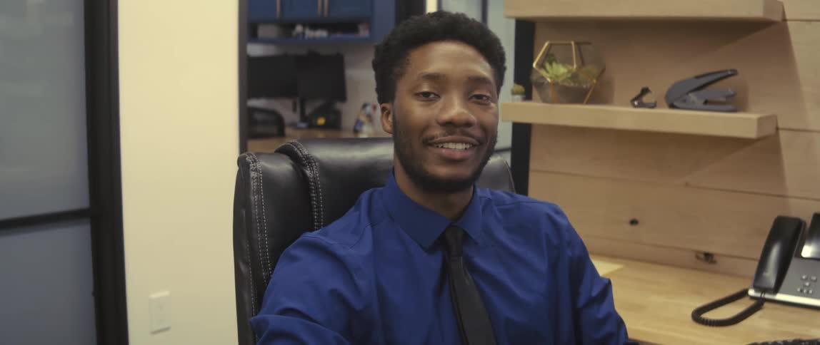 A man in a blue dress shirt sitting in an office chair looking straight ahead and then he turns around and starts smiling to the camera