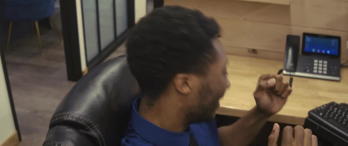 A young man in the blue dress shirt is in an office sitting on a chair and he is celebrating and then he turns the chair and starts typing on the keyboard