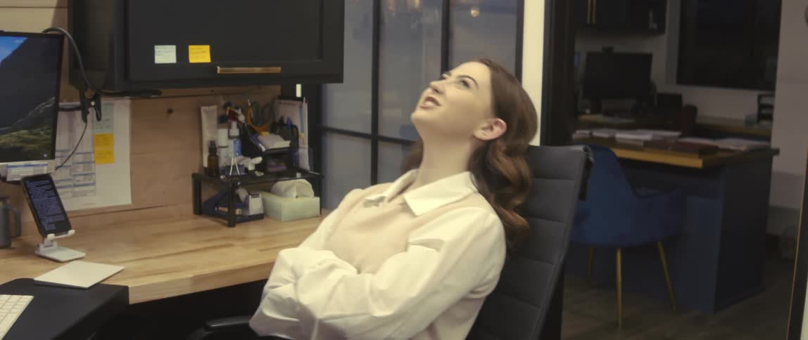 A relaxed office worker wearing a white shirt reclines on a chair in her cubicle