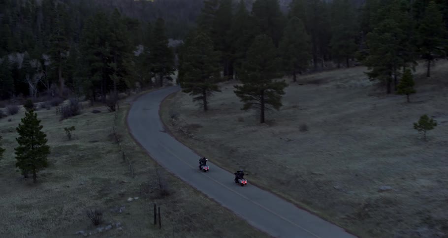 Two motorcycles are being driven down a road that sits between heavily wooded areas