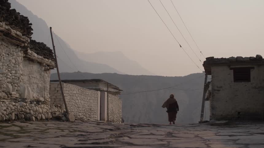 A robed person walks down a cobblestone street lined with stone buildings and mountains visible in the background