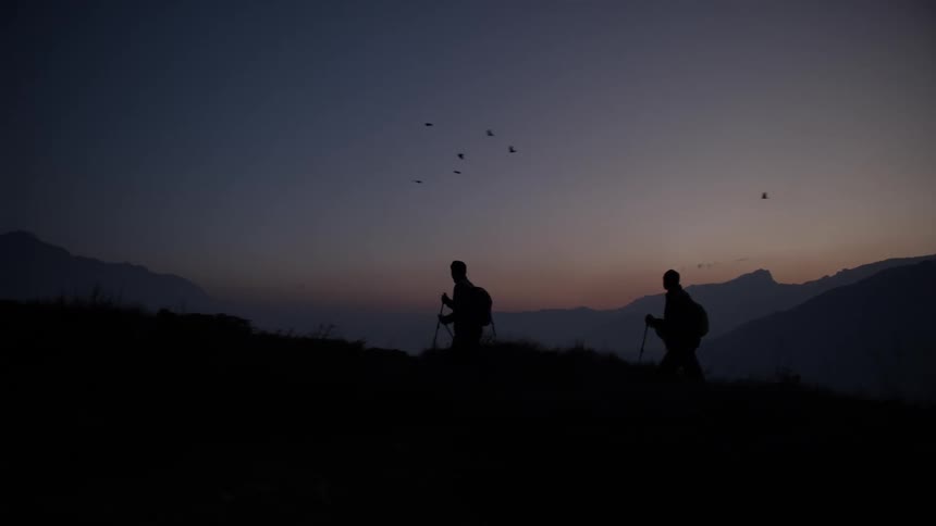 Two backpackers hike along a slope overlooking a foggy valley while birds fly through the red tinted sky at night