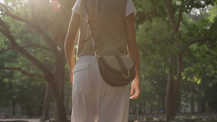 A woman with a purse walks slowly through a forest