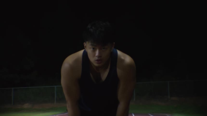 Man in tank top with a look of determination resting his hands on his legs while standing on a running track at night