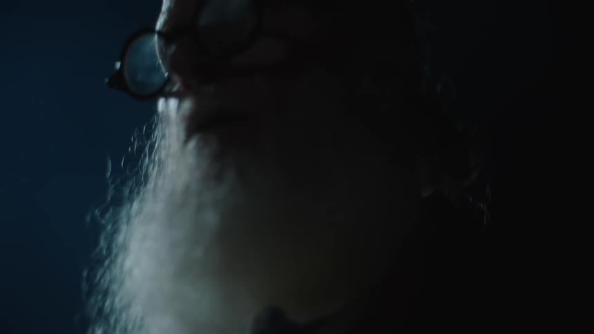 An elderly man with a large white beard and reading glasses is shaking his head back and forth in an outdoor setting at night