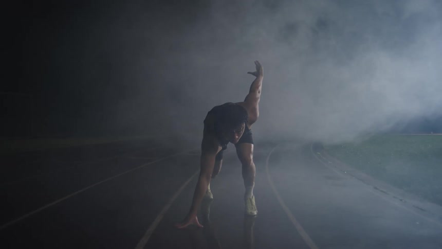 A runner in athletic gear crouches in a starting position on a misty track ready to launch forward under the dimly lit atmosphere