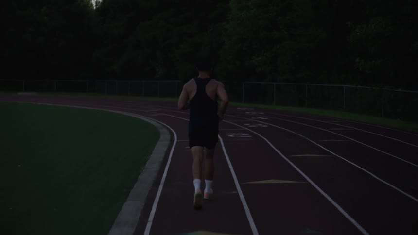 A man is facing away and running on a red painted race track and he is approaching a curve