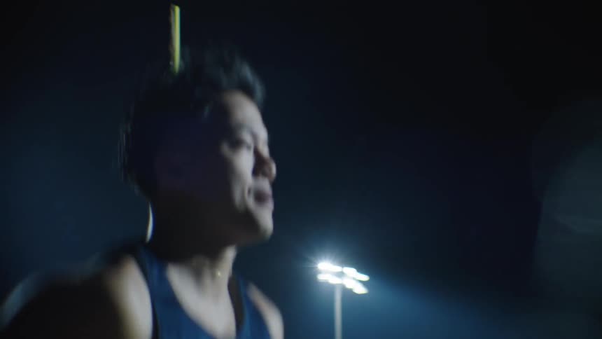 A man is wearing a tank top and is running in an outdoor environment at night illuminated by spotlights