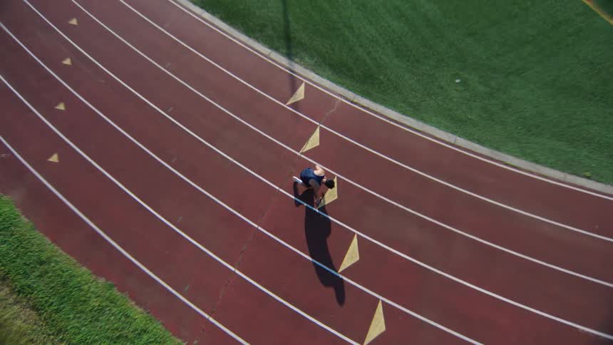 A man in a blue tank top is preparing to run on a red running track seen from above