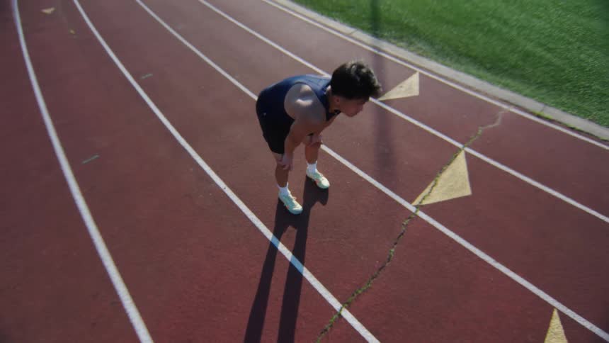 A man is on a running track standing and then puts his hands on his knee he is looking tired