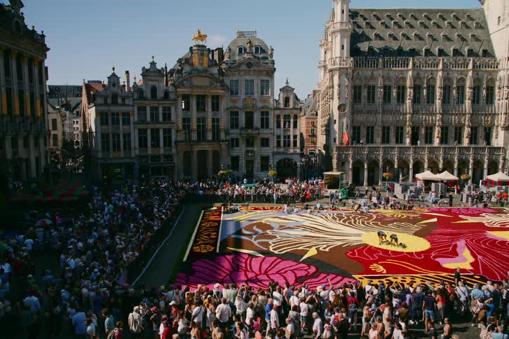 A large crowd of people gather near a city square near a large piece of artwork covering a large part of the square