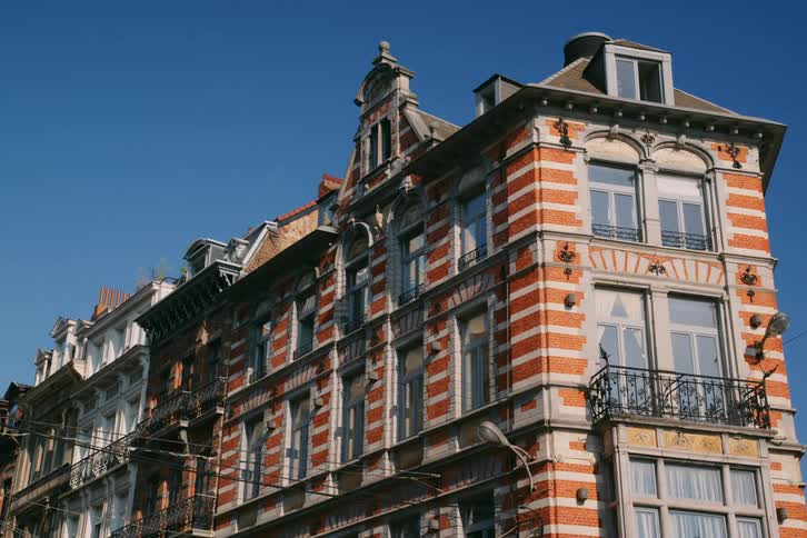A row of historic brick buildings with ornate facades and balconies bathed in bright sunlight against a clear blue sky