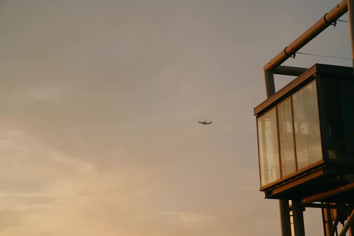 A small rectangular building with a glass facade against a sky with a setting sun and a small airplane flying overhead