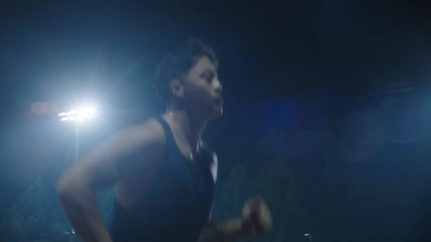 A young man in a tank top is running in an outdoor environment at night illuminated by some spotlights