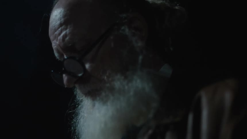 An old man with a large beard and glasses looks around outdoors at night