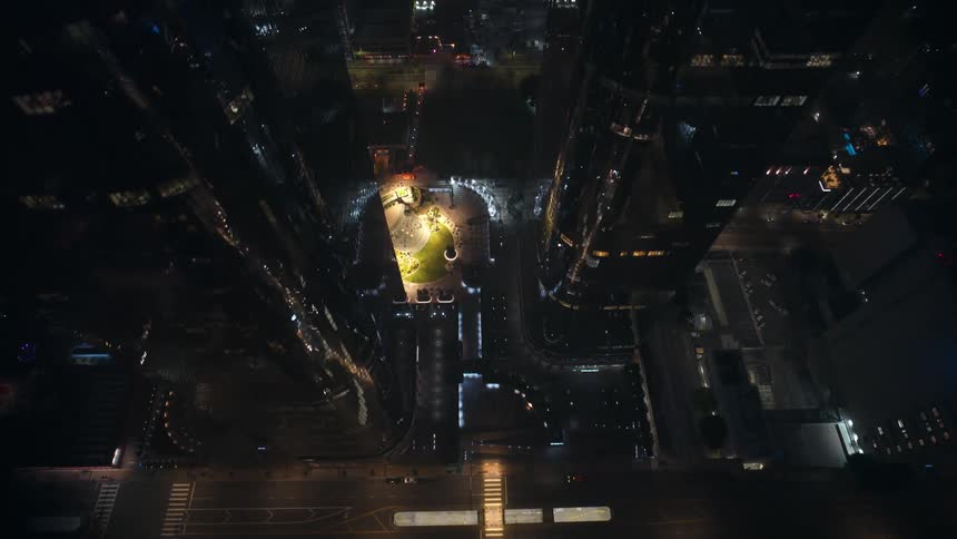 An aerial fly over of a metropolitan city center with tall buildings and a lit up square at night